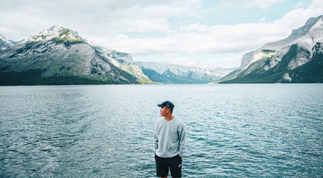 a man in gray sweater standing near the body of water while looking at the mountain