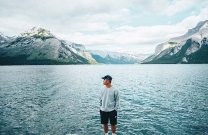 a man in gray sweater standing near the body of water while looking at the mountain